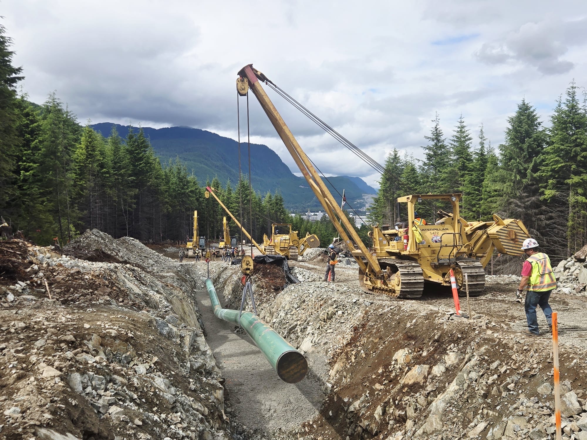 Yellow cranes lower first length of natural gas pipeline into trench in northern B.C. while workers in PPE supervise along the edge of the trench.