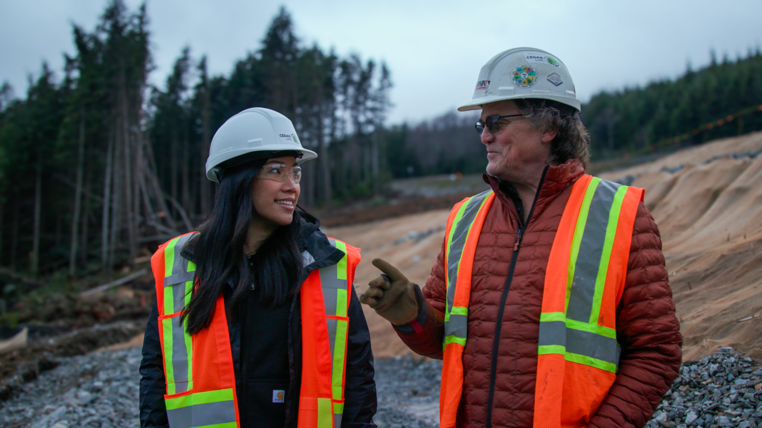 A younger woman with dark hair and an older man with grey hair walk towards camera in discussion along proposed pipeline right of way wearing orange safety vests and white helmets.